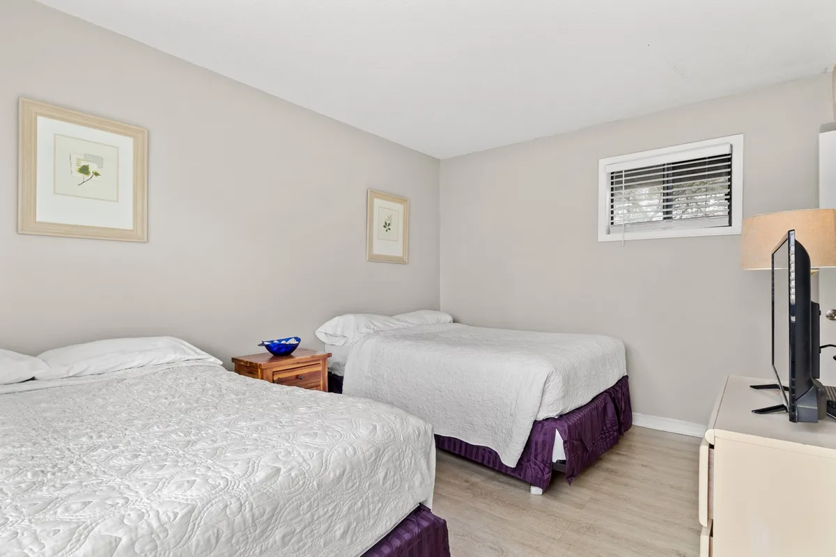 The second bedroom offers two beds with crisp white linens, separated by a wooden nightstand. The decor is tranquil, with a decorative blue bowl and framed wall art, creating a peaceful retreat. The room has a clean, minimalist look ideal for shared accommodation.