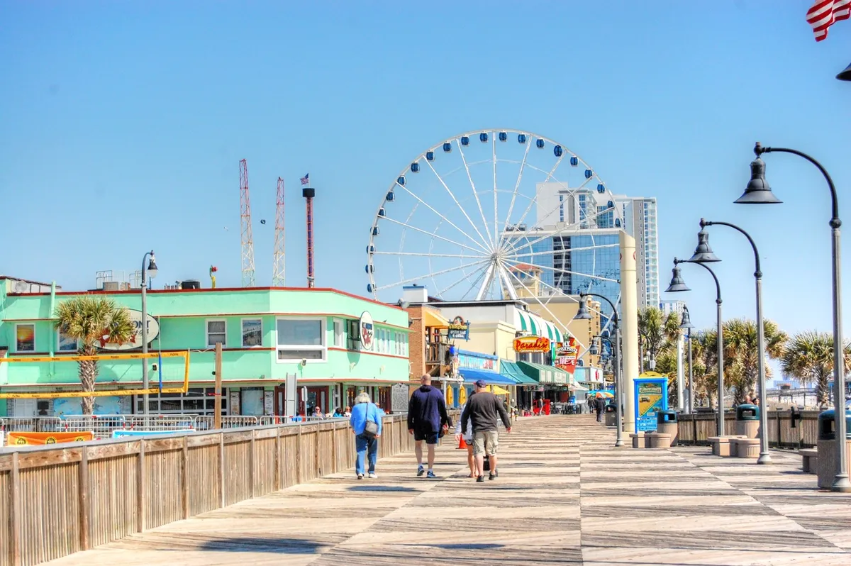 A lively boardwalk scene featuring a towering Ferris wheel, colorful buildings, and people strolling amidst a festive coastal atmosphere. The scene conveys the energy and variety of Myrtle Beach’s attractions, from amusement rides to local shops and snacks.