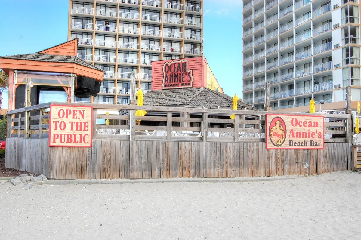 This image showcases the iconic Ocean Annie's Beach Bar, with its recognizable sign, lively yellow umbrellas, and open wooden structure. The bar looks inviting, set just steps from the sand and perfectly situated for groups looking to enjoy live music, tropical drinks, and the vibrant beach atmosphere.