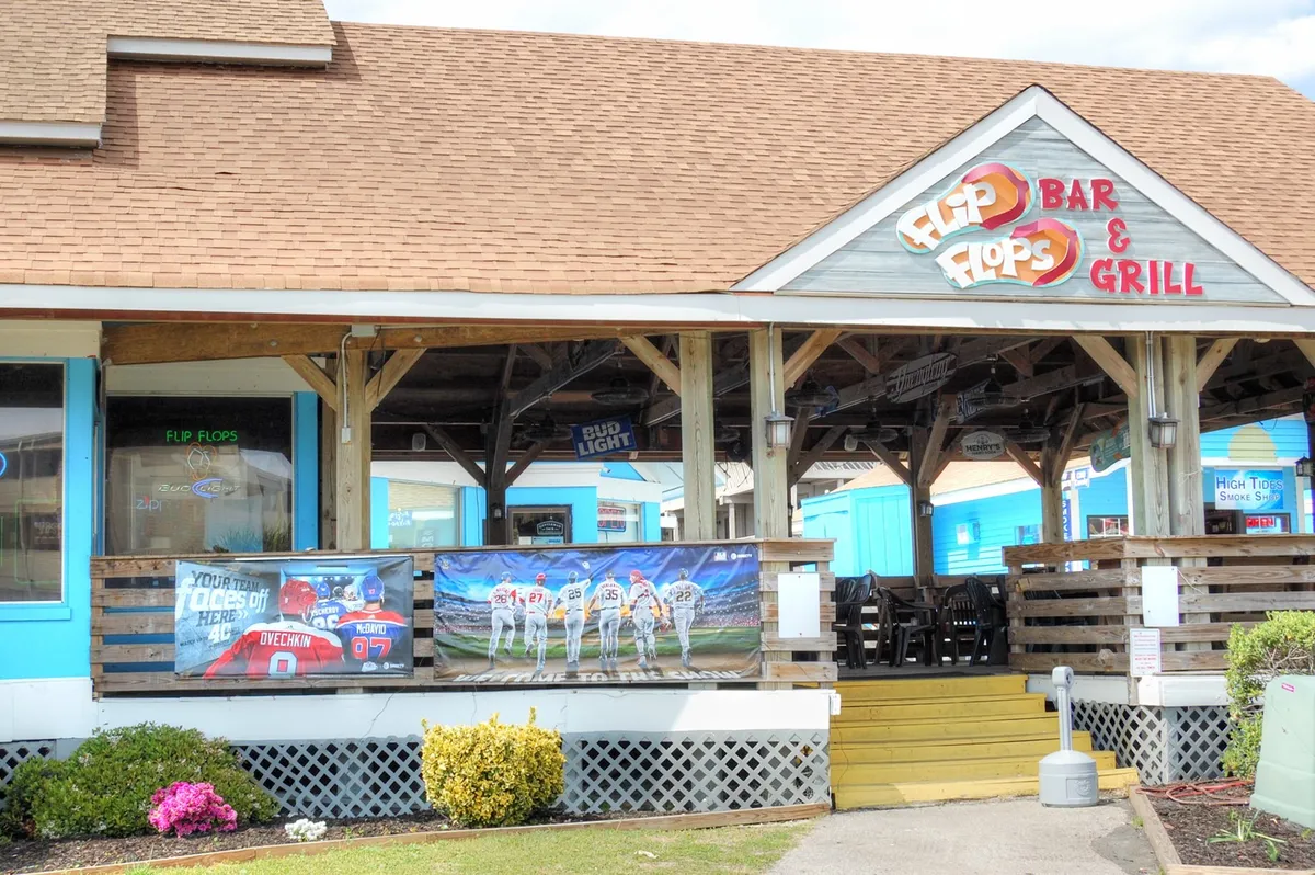A welcoming outdoor dining area with the prominent signage of 'Flip Flops Bar & Grill,' decorated with sports banners and featuring plenty of space for socializing and enjoying meals with friends.