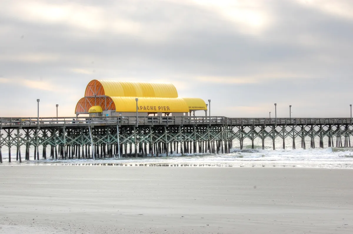 This image captures the striking Apache Pier jutting out into the ocean, with gentle waves lapping beneath and the pier building anchoring the scene. The focus is on the classic structure, set against a backdrop of blue water and open sky.