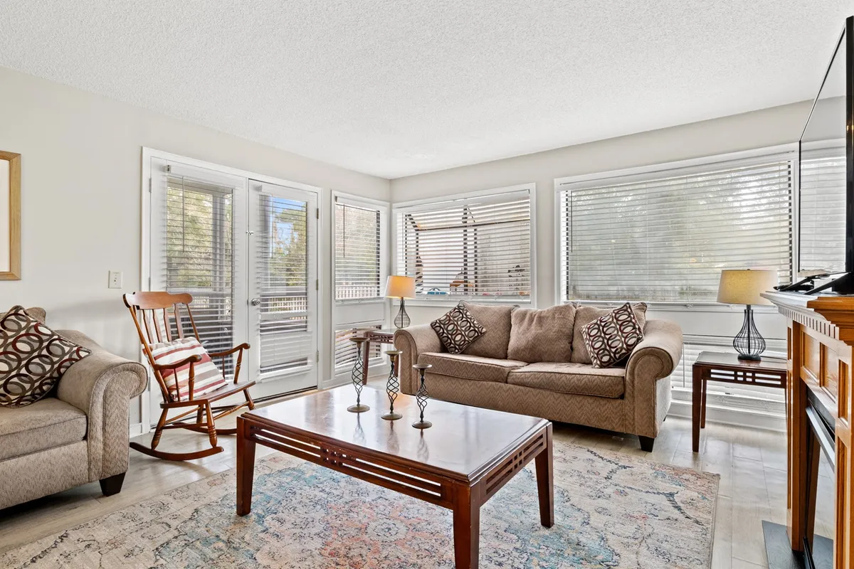 The image showcases a bright and inviting living room, featuring a cozy sofa, a classic rocking chair by large sunlit windows, and a central coffee table perfect for games or morning coffee. The space exudes comfort and warmth, with natural light streaming in to create a relaxing coastal ambiance.