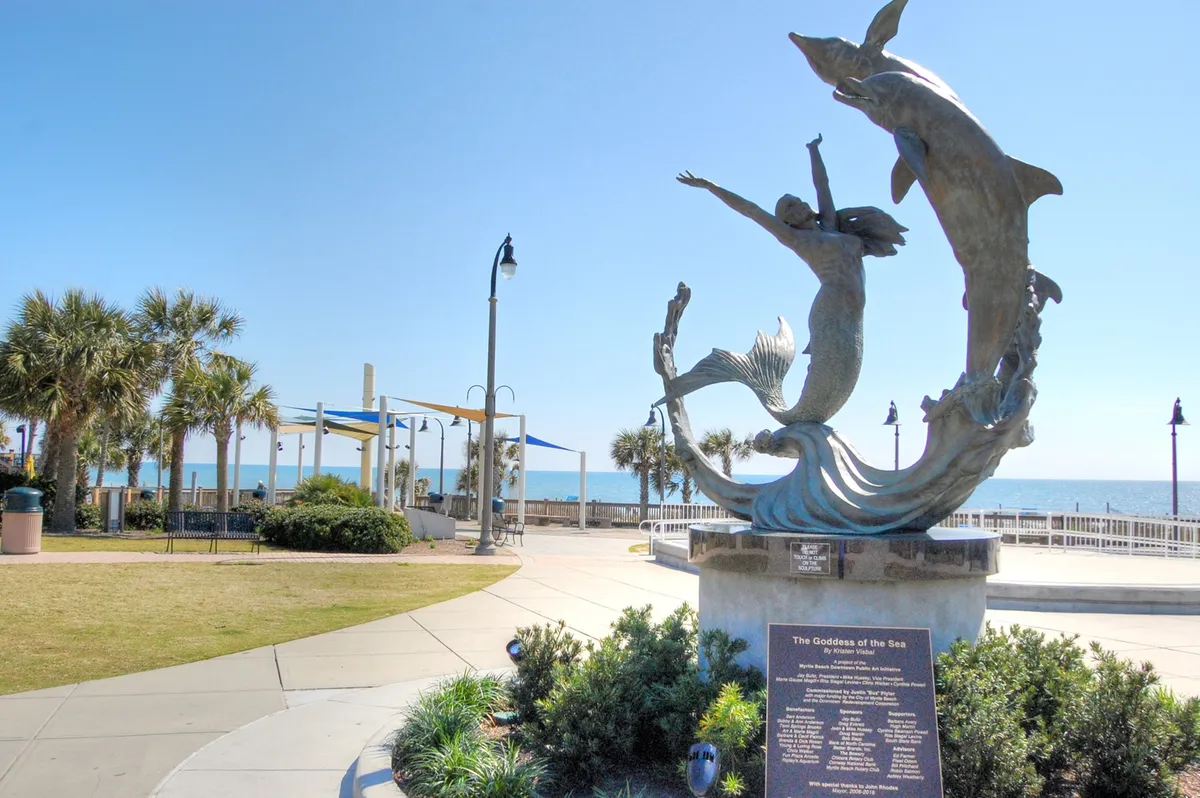 This exterior image showcases a serene oceanfront scene featuring a statue of a goddess and dolphins, framed by palm trees and a curving walkway. The ocean is visible in the background, and the setting sun casts a warm glow, evoking a tranquil, romantic seaside atmosphere.