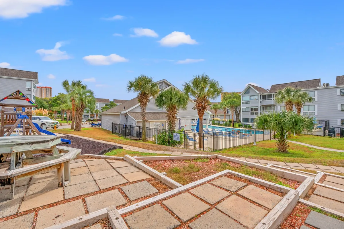 A vibrant exterior scene showcasing a large swimming pool surrounded by palm trees, with children enjoying the playground and people relaxing outdoors. The apartment buildings are visible in the background, creating a lively and communal atmosphere.