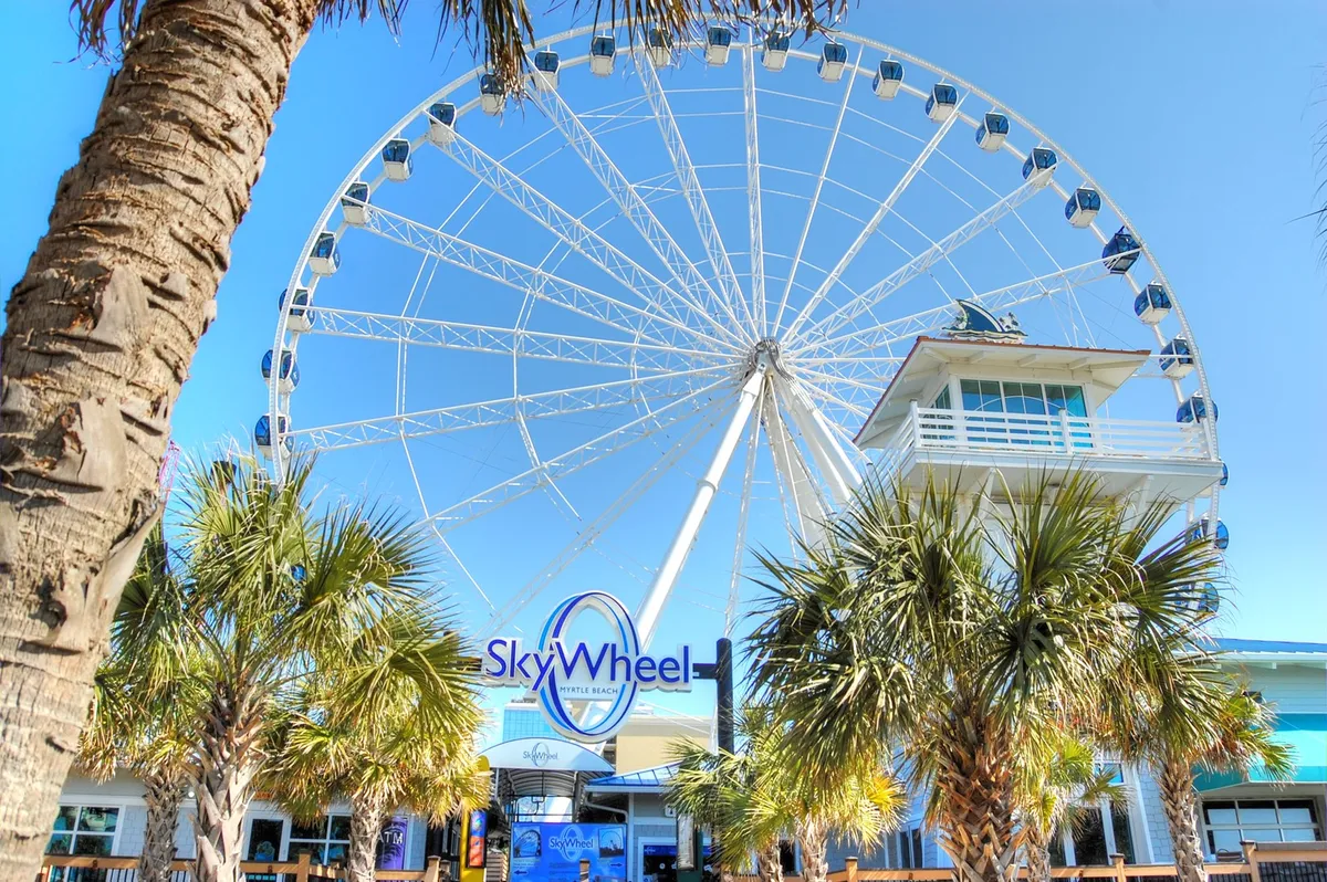 This exterior shot captures the SkyWheel towering over the Myrtle Beach boardwalk, framed by palm trees and an inviting entrance. The scene is filled with vacationers enjoying the ride and the scenic oceanfront setting, evoking excitement and adventure.