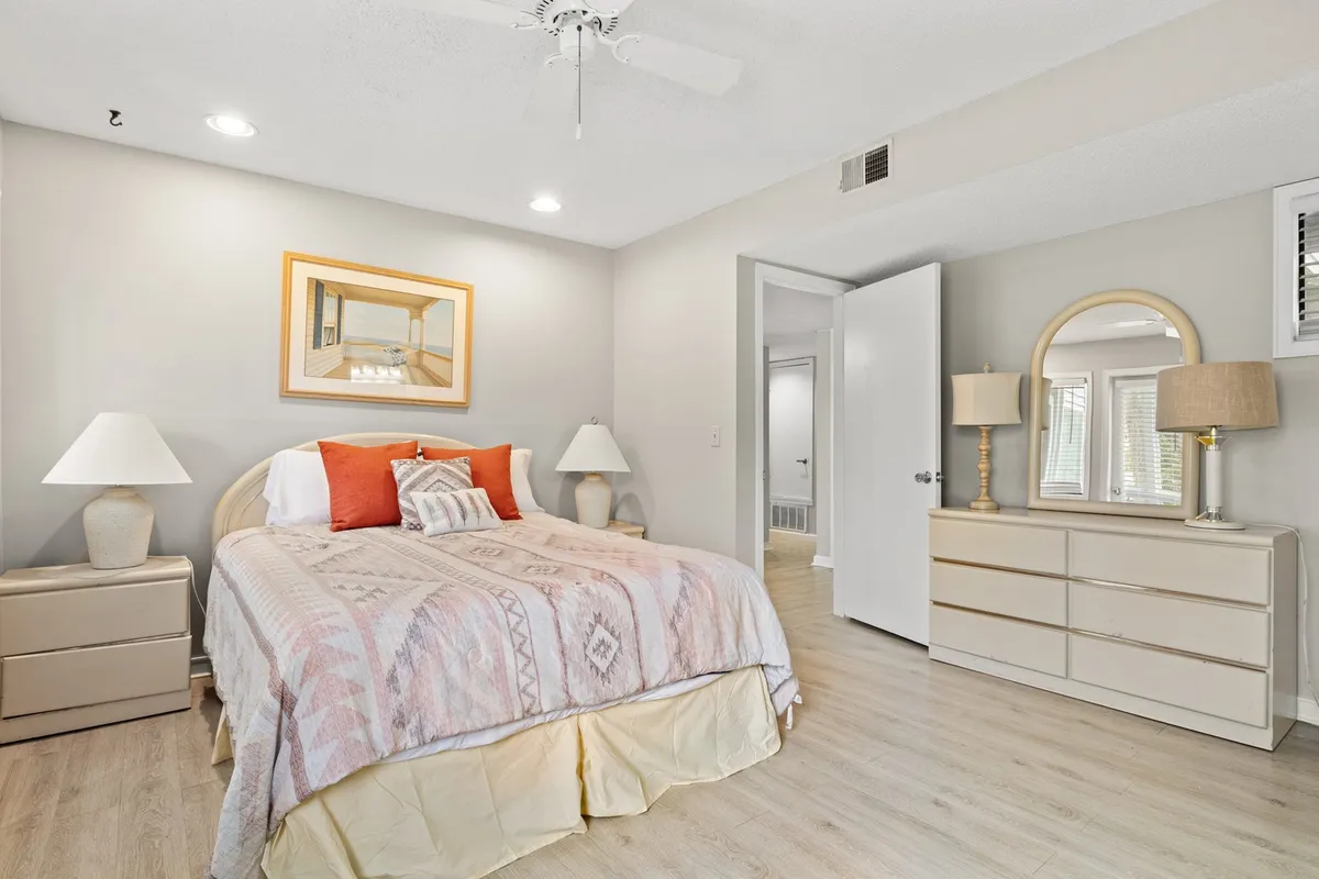 A cozy bedroom featuring a neatly made bed with decorative pillows, flanked by nightstands with lamps for ambient lighting. A mirror rests on the dresser, and framed artwork adds a thoughtful touch to the decor. Sunlight streams through the window, creating a bright and peaceful environment.